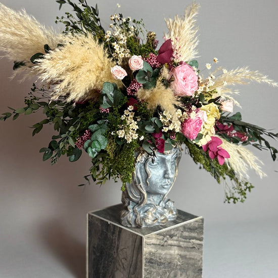 Arreglo floral con plumas de pampas y rosas en jarrón de rostro sobre pedestal de mármol