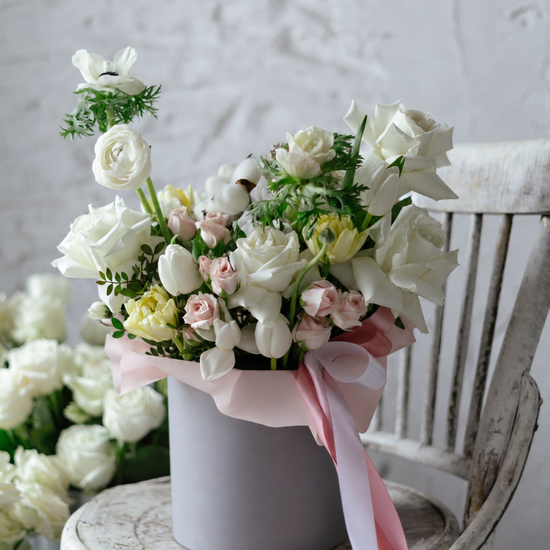 Caja gris con rosas blancas, mini rosas rosa palo, tulipanes amarillos, anémonas y algodón, con cintas rosas sobre silla de madera