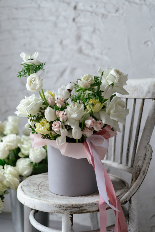 Caja gris con rosas blancas, mini rosas rosa palo, tulipanes amarillos, anémonas y algodón, con cintas rosas sobre silla de madera