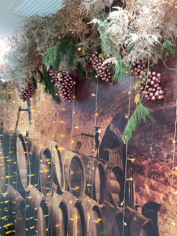 Decoración navideña con racimos de uvas artificiales y luces colgantes sobre un mural de barricas de vino
