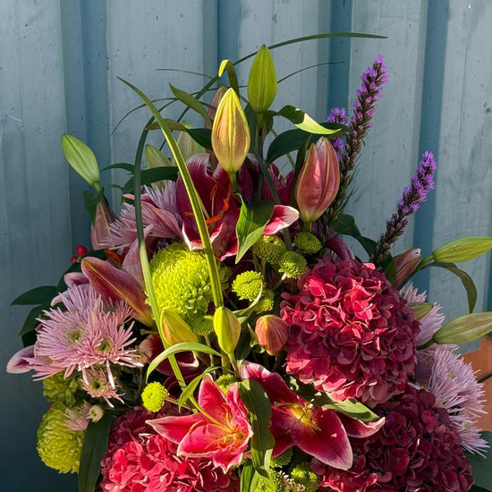 Centro floral con hortensias granates, lilium rosa, crisantemos verdes y lilas y liatris morado en maceta blanca
