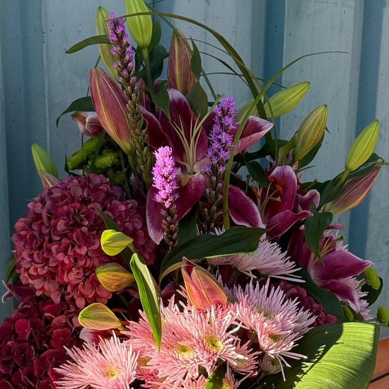 Centro floral con lilium, liatris morado, hortensia granate y crisantemos rosas rodeados de hojas verdes enrolladas