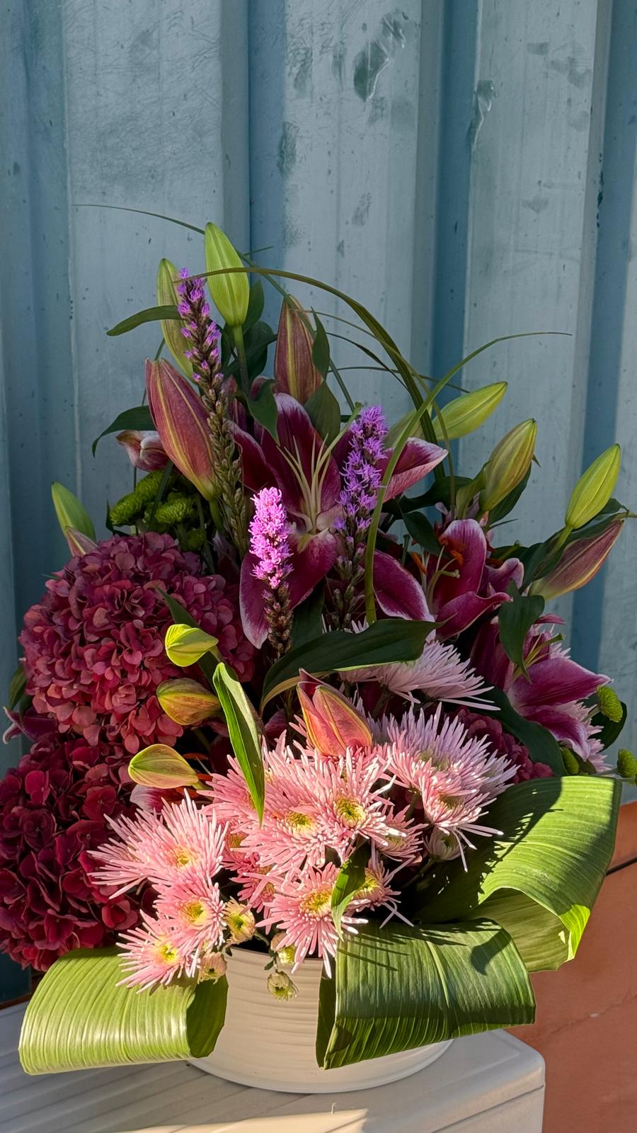 Centro floral con lilium, liatris morado, hortensia granate y crisantemos rosas rodeados de hojas verdes enrolladas