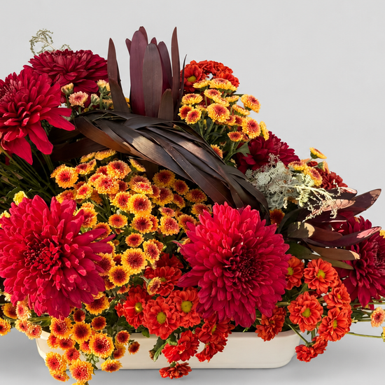 Centro de mesa bajo con crisantemos rojos, naranjas y amarillos, leucadendron y hojas trenzadas en bandeja blanca