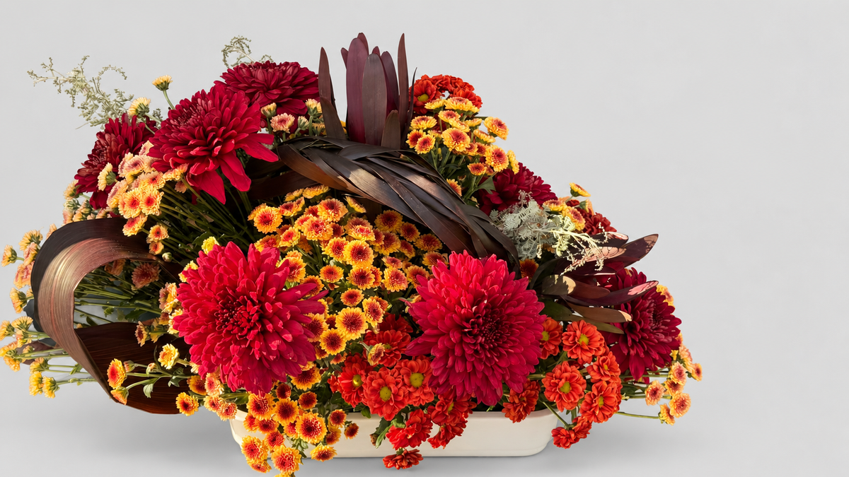 Centro de mesa bajo con crisantemos rojos, naranjas y amarillos, leucadendron y hojas trenzadas en bandeja blanca