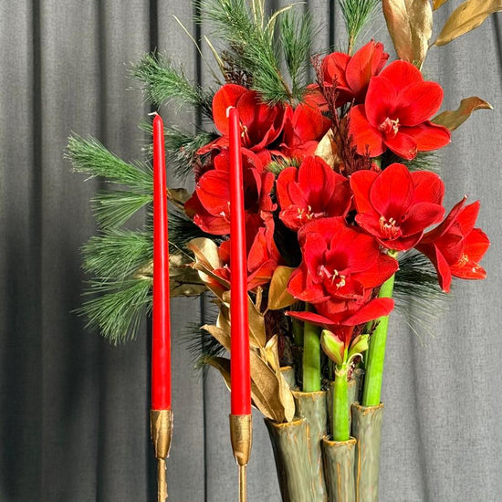 Centro navideño de amarilis rojas con pino y hojas doradas en jarrón de cerámica verde, junto a velas rojas, ANHUÁ Flowers Murcia