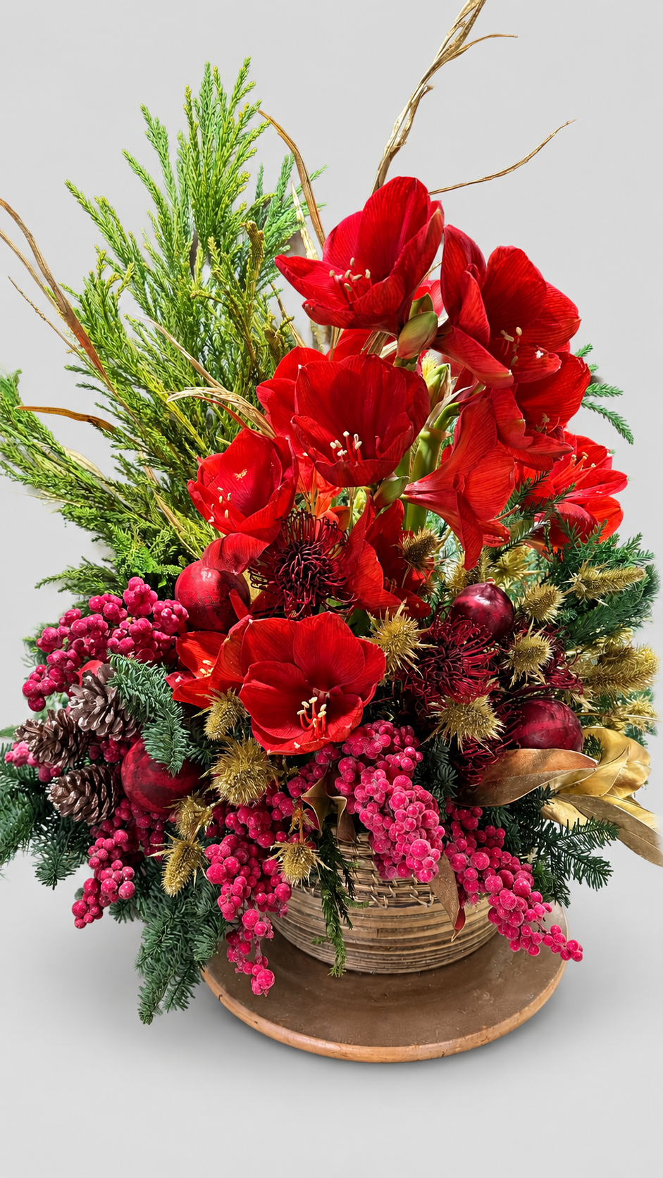 Centro navideño en cesta con amarilis rojas, protea, cardos dorados, piñas, bayas rosas y ramas de pino