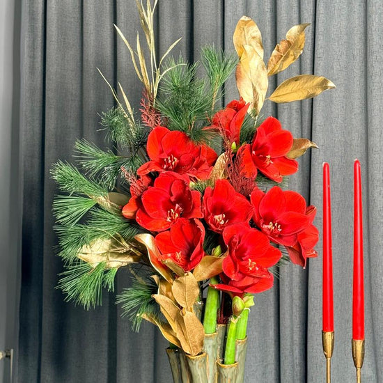 Composición de Navidad con amarilis rojas, pino y hojas doradas en jarrón de cerámica sobre mesa de madera con velas rojas