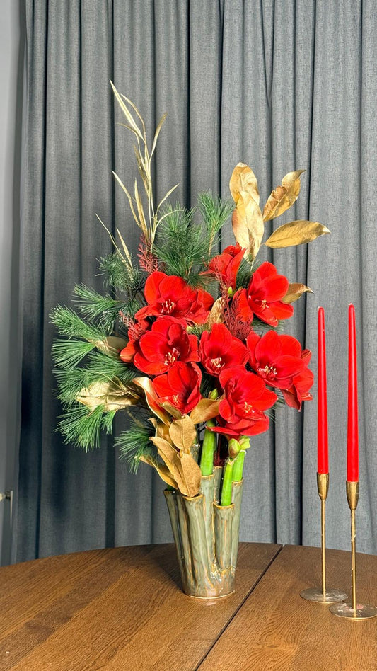 Composición de Navidad con amarilis rojas, pino y hojas doradas en jarrón de cerámica sobre mesa de madera con velas rojas