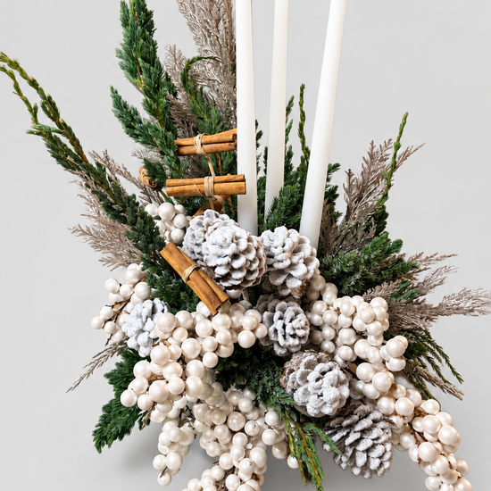 Composición navideña con tres velas blancas, piñas nevadas, ramas de canela, bayas perladas y verdes de abeto
