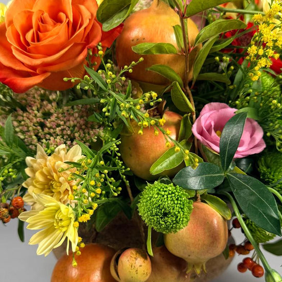Composición otoñal en copa dorada con granadas, rosas naranjas, lisianthus rosa y crisantemos amarillos