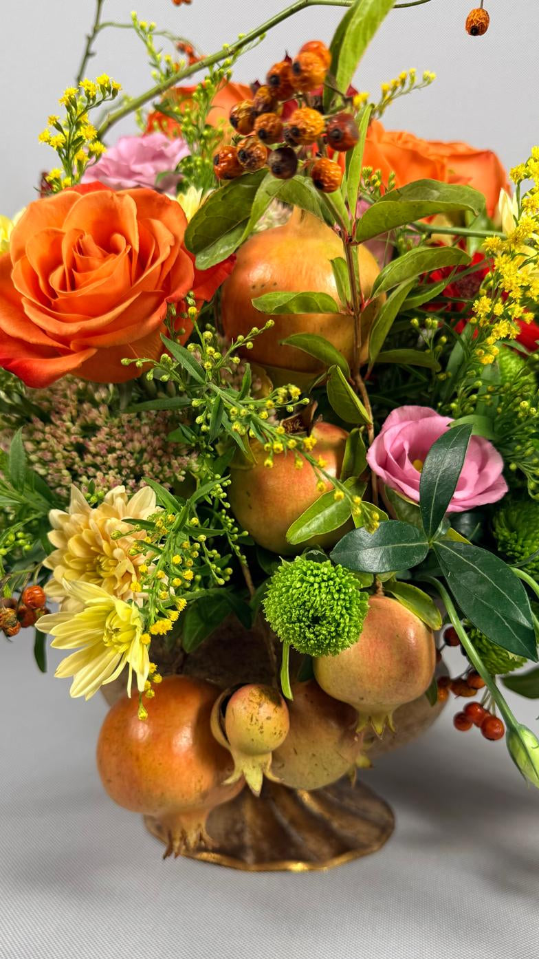 Composición otoñal en copa dorada con granadas, rosas naranjas, lisianthus rosa y crisantemos amarillos
