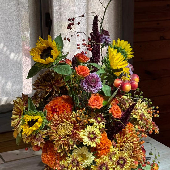 Composición otoñal con girasoles, rosas naranjas, crisantemos, celosía, amaranto, escabiosas y manzanas ornamentales junto a una ventana