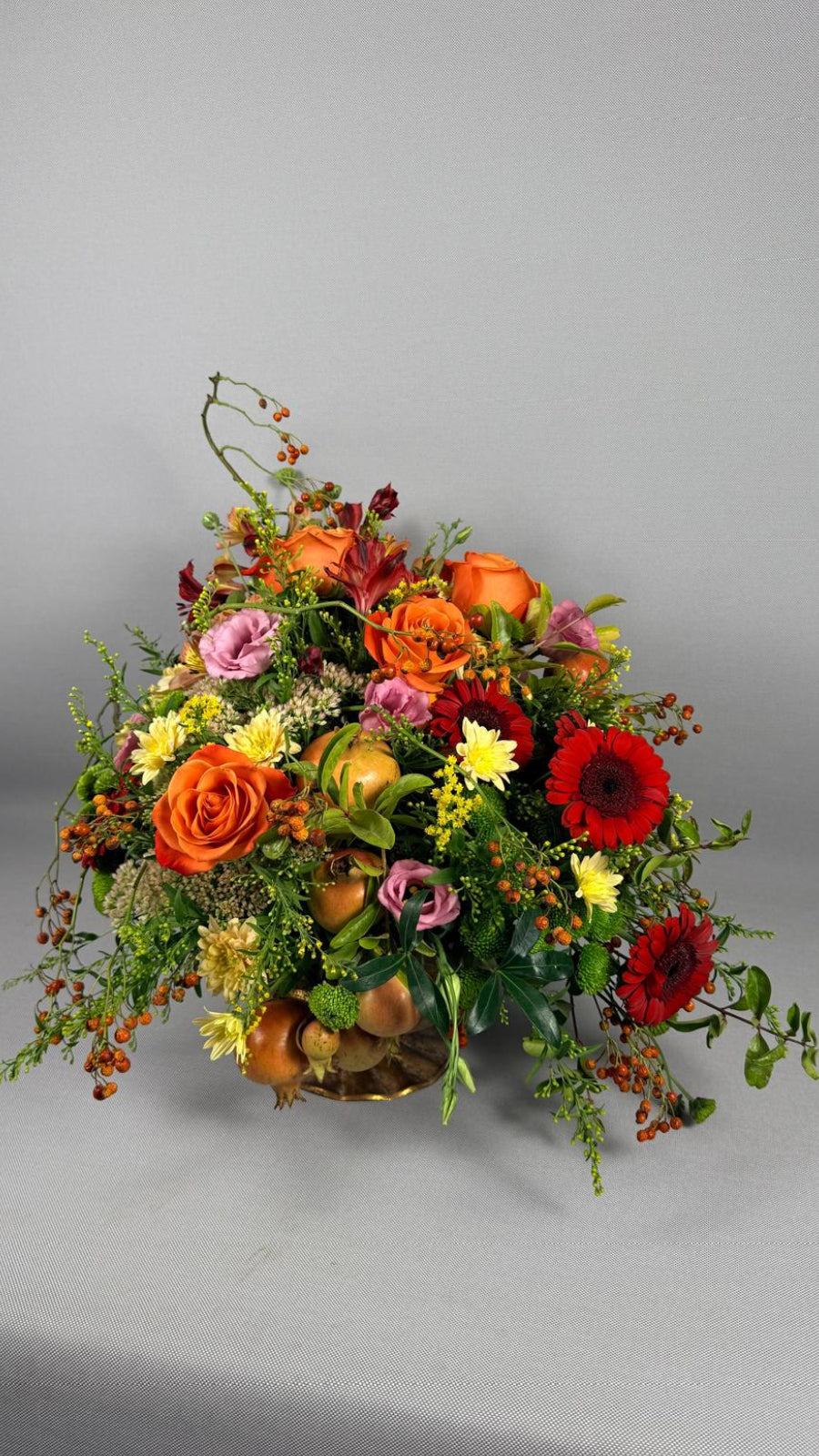 Composición floral otoñal con rosas naranjas, gerberas rojas, lisianthus rosa, crisantemos amarillos y granadas