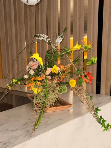 Arreglo floral con calas amarillas y flores naranjas sobre una base de madera y fibras naturales