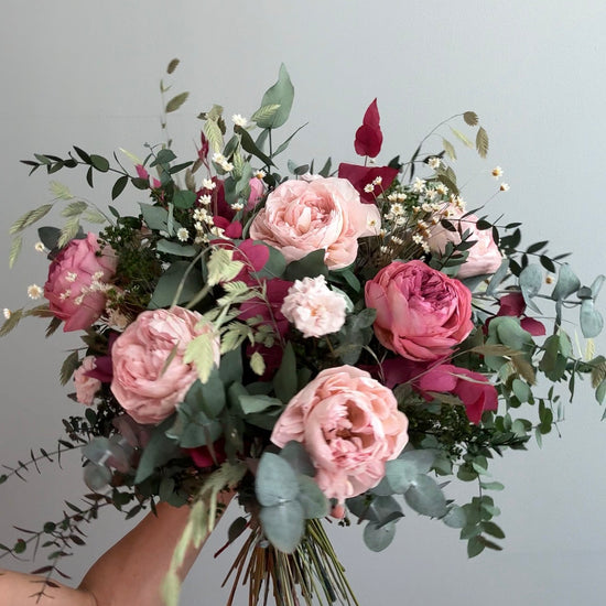 Mano sosteniendo ramo de rosas rosas y coral con eucalipto sobre fondo gris