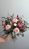 Mano sosteniendo ramo de rosas rosas y coral con eucalipto sobre fondo gris
