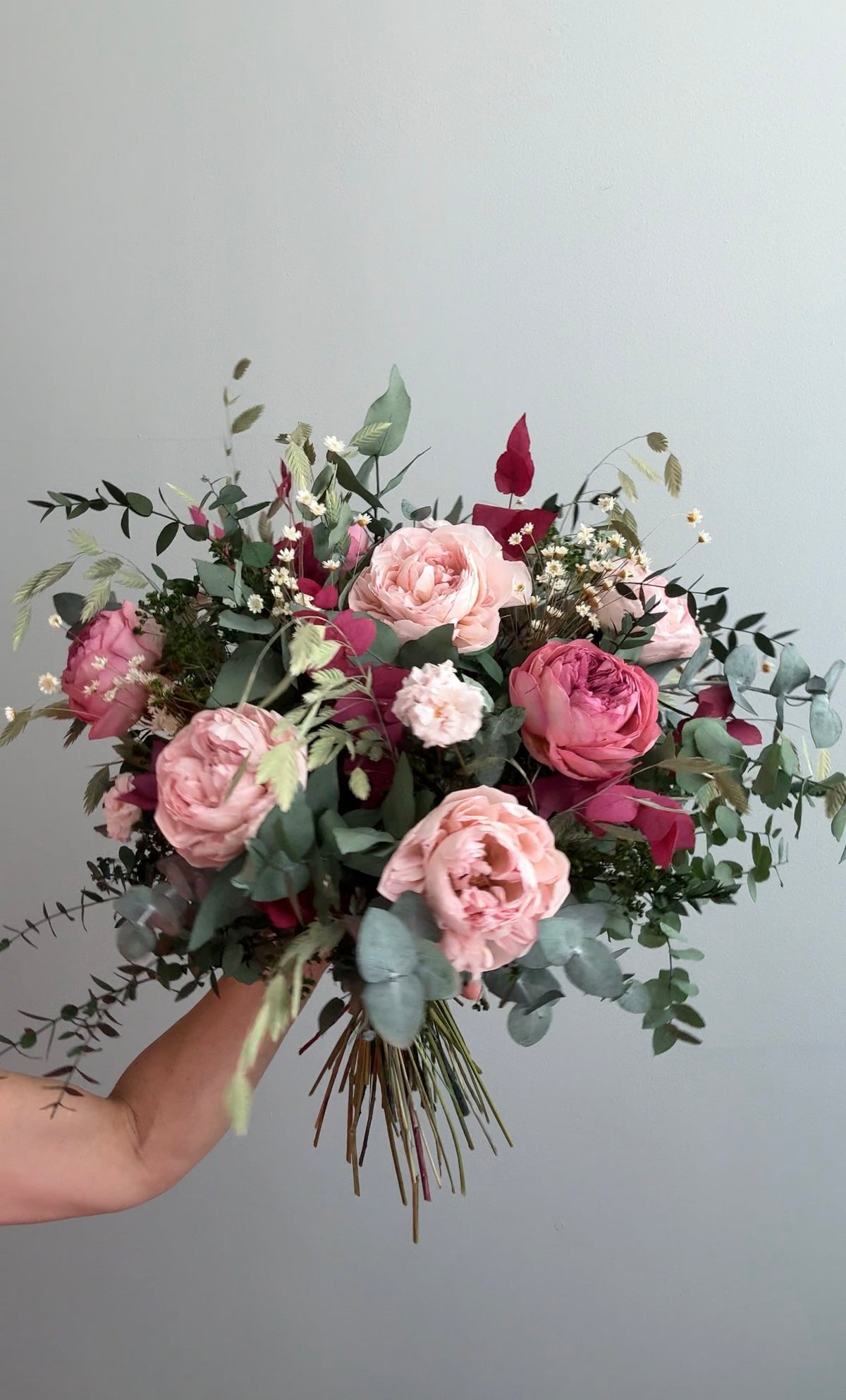 Mano sosteniendo ramo de rosas rosas y coral con eucalipto sobre fondo gris