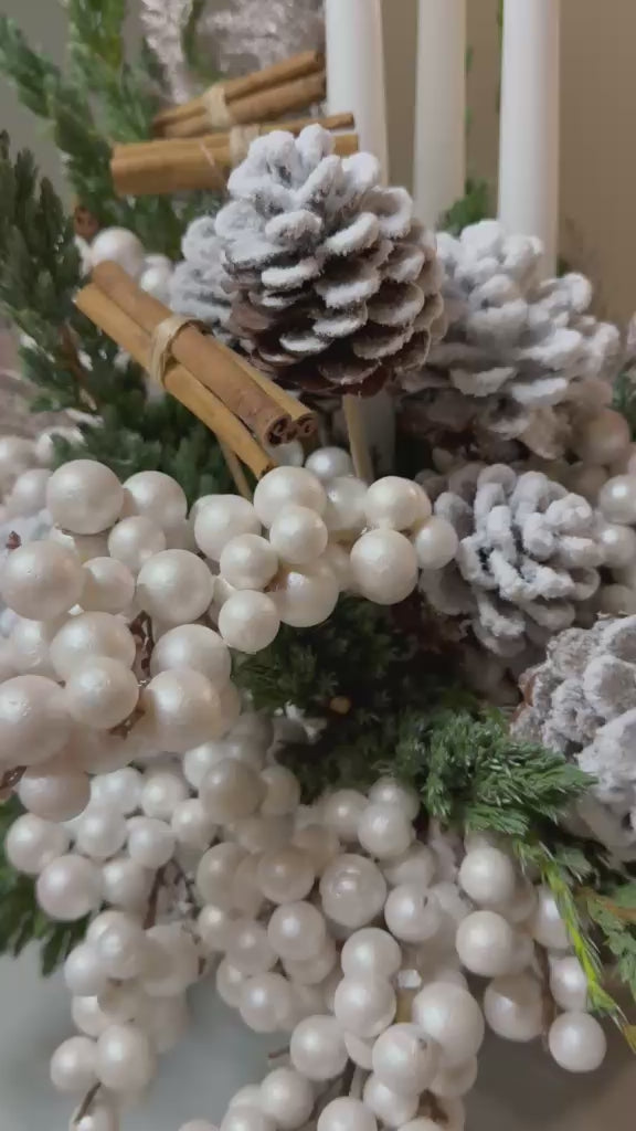 Composición navideña con piñas nevadas, canela, bayas perladas, verdes de abeto y velas blancas