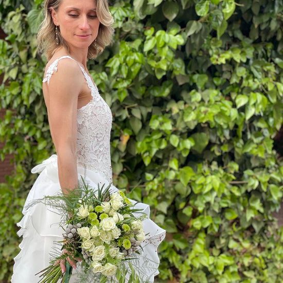 Novia con vestido de encaje y ramo de rosas blancas y crisantemos verdes frente a un muro de hiedra