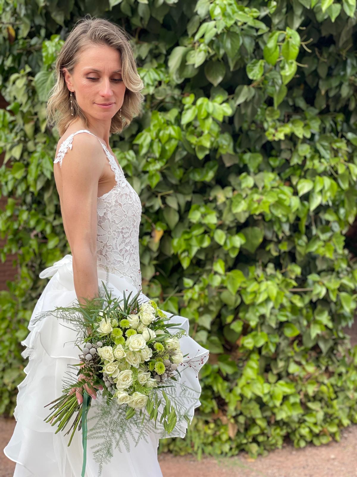 Novia con vestido de encaje y ramo de rosas blancas y crisantemos verdes frente a un muro de hiedra