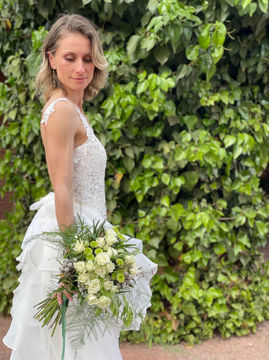 Novia con vestido de encaje y ramo de rosas blancas y crisantemos verdes frente a un muro de hiedra