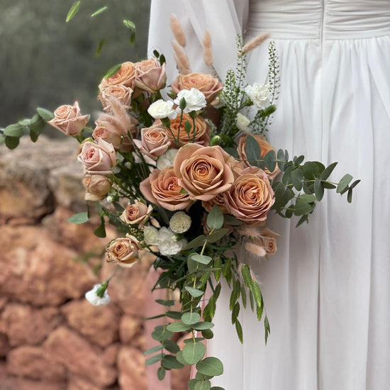 Ramo de novia con rosas capuchino, claveles blancos, lagurus y eucalipto colgante junto a un muro de piedra