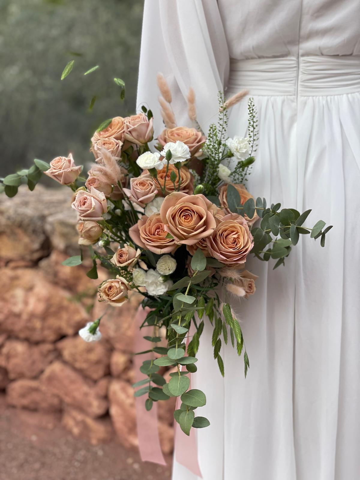 Ramo de novia con rosas capuchino, claveles blancos, lagurus y eucalipto colgante junto a un muro de piedra