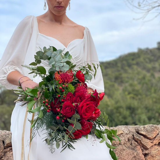 Novia sentada con ramo de rosas rojas y leucospermum atado con cinta dorada, montañas al fondo