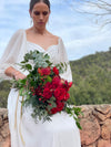 Novia sentada con ramo de rosas rojas y leucospermum atado con cinta dorada, montañas al fondo