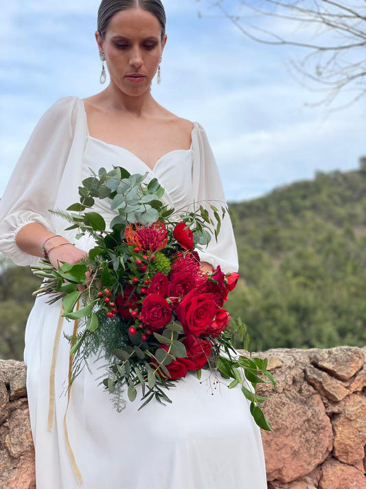 Novia sentada con ramo de rosas rojas y leucospermum atado con cinta dorada, montañas al fondo