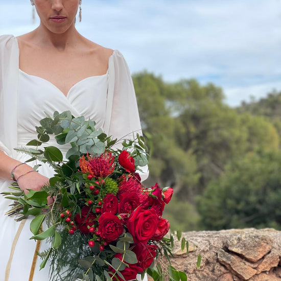 Novia al aire libre sosteniendo un ramo de novia de rosas rojas, leucospermum, hypericum y eucalipto