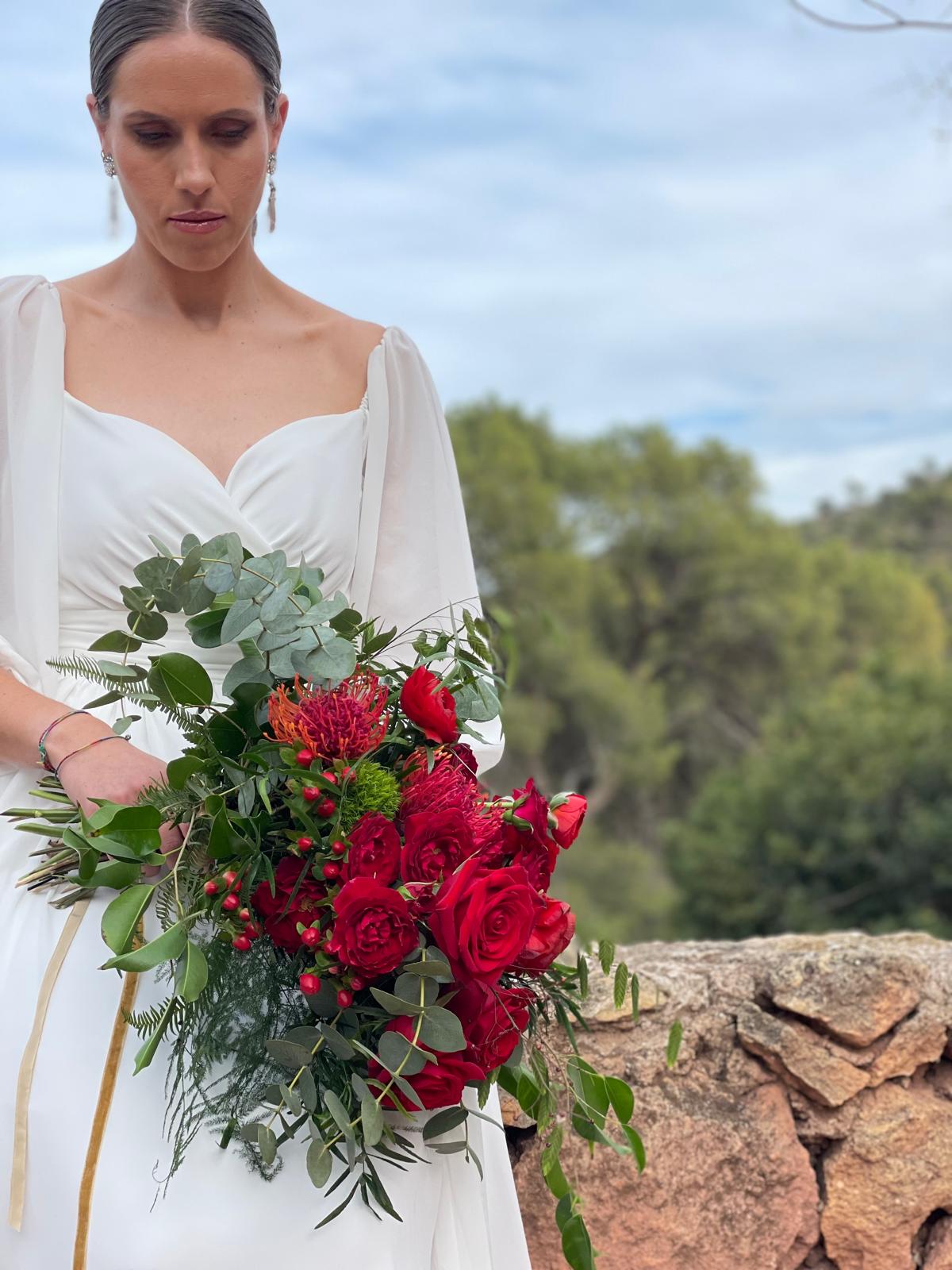 Novia al aire libre sosteniendo un ramo de novia de rosas rojas, leucospermum, hypericum y eucalipto