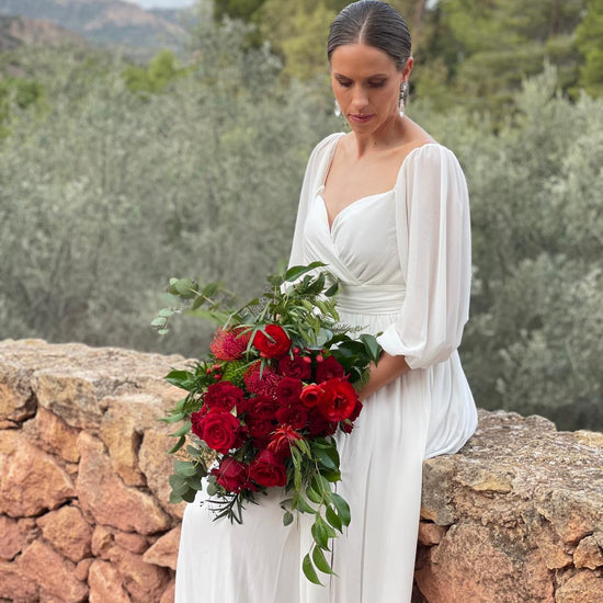 Novia sentada sobre un muro de piedra con ramo de novia de rosas rojas, leucospermum y eucalipto