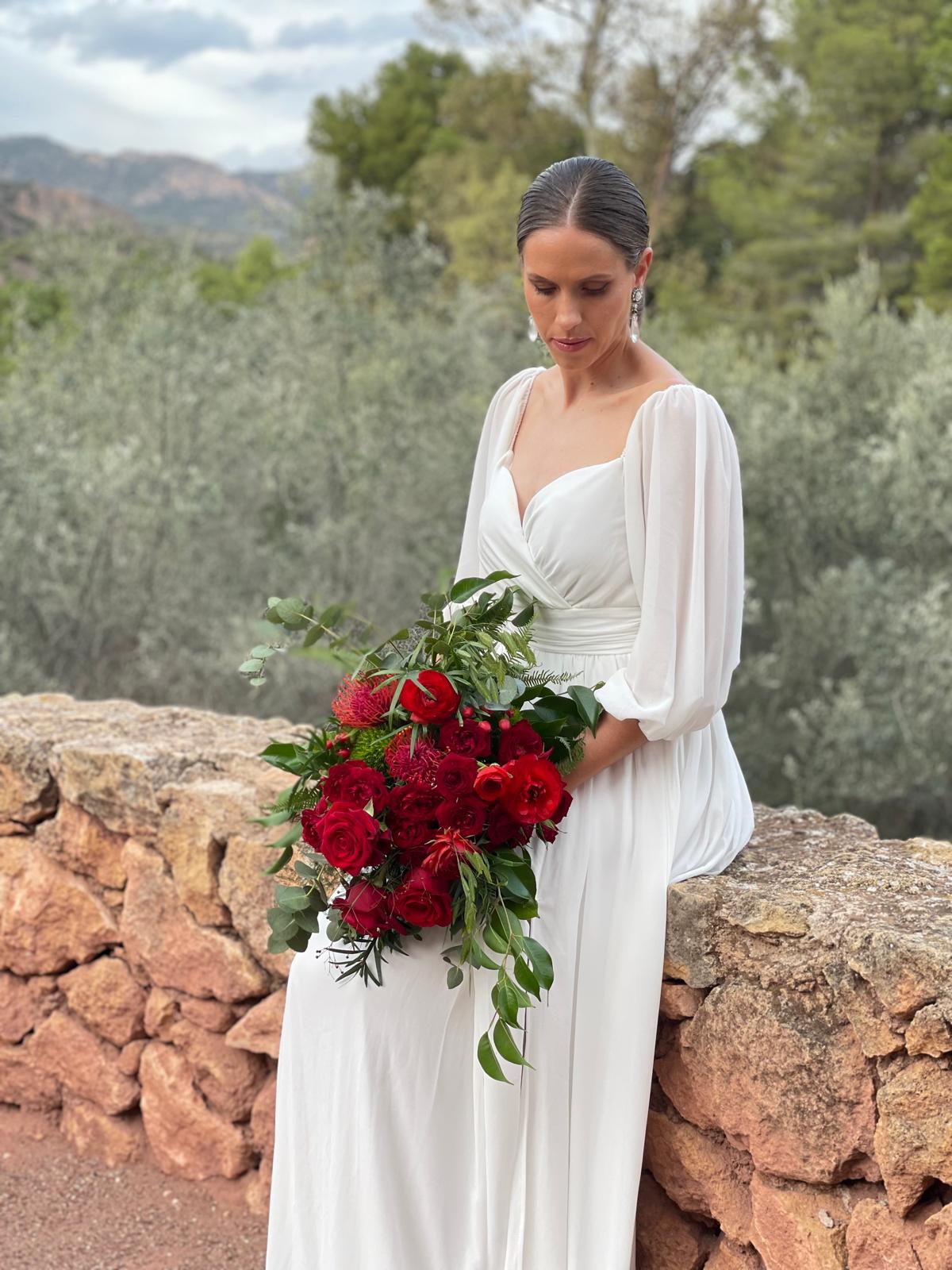 Novia sentada sobre un muro de piedra con ramo de novia de rosas rojas, leucospermum y eucalipto