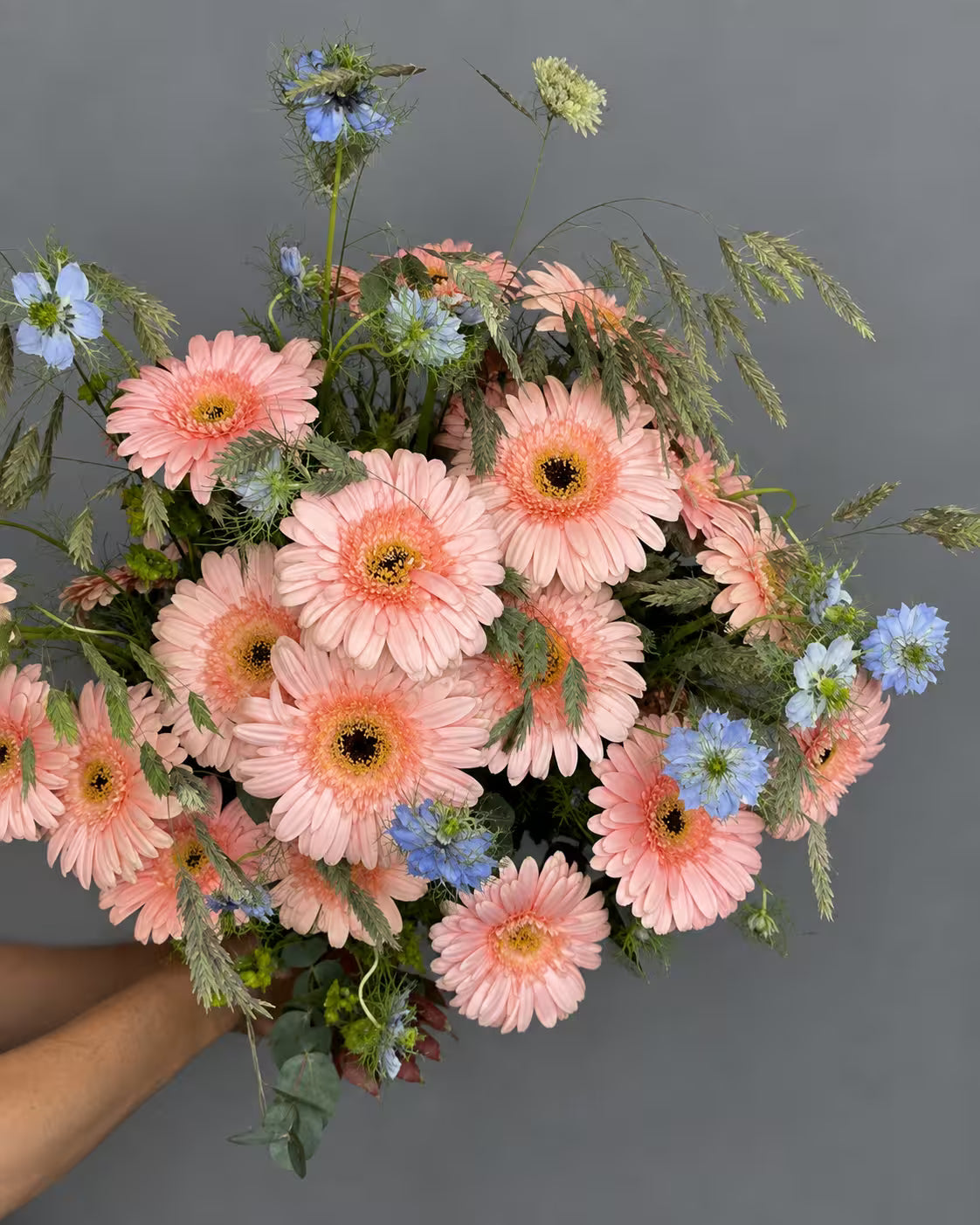 Ramo de gerberas color melocotón con nigella azul, espigas y eucalipto, sostenido en la mano