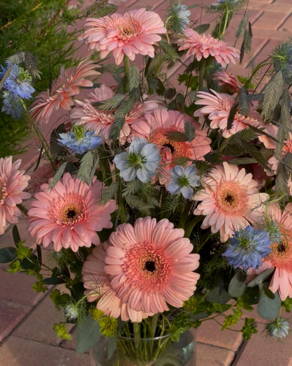 Ramo de gerberas rosas con nigella azul y espigas en jarrón de cristal, fotografiado al aire libre