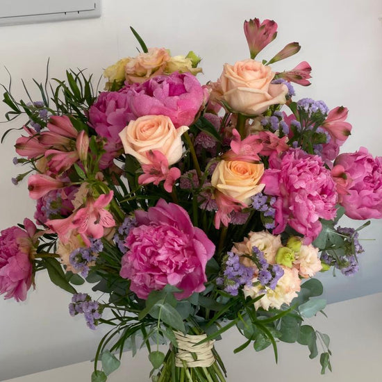 Ramo de peonías fucsia, rosas melocotón, alstroemerias y limonium lila con eucalipto, atado con rafia