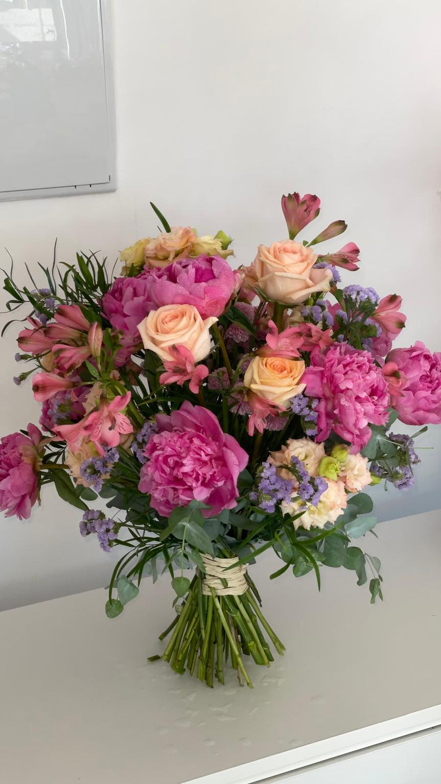 Ramo de peonías fucsia, rosas melocotón, alstroemerias y limonium lila con eucalipto, atado con rafia