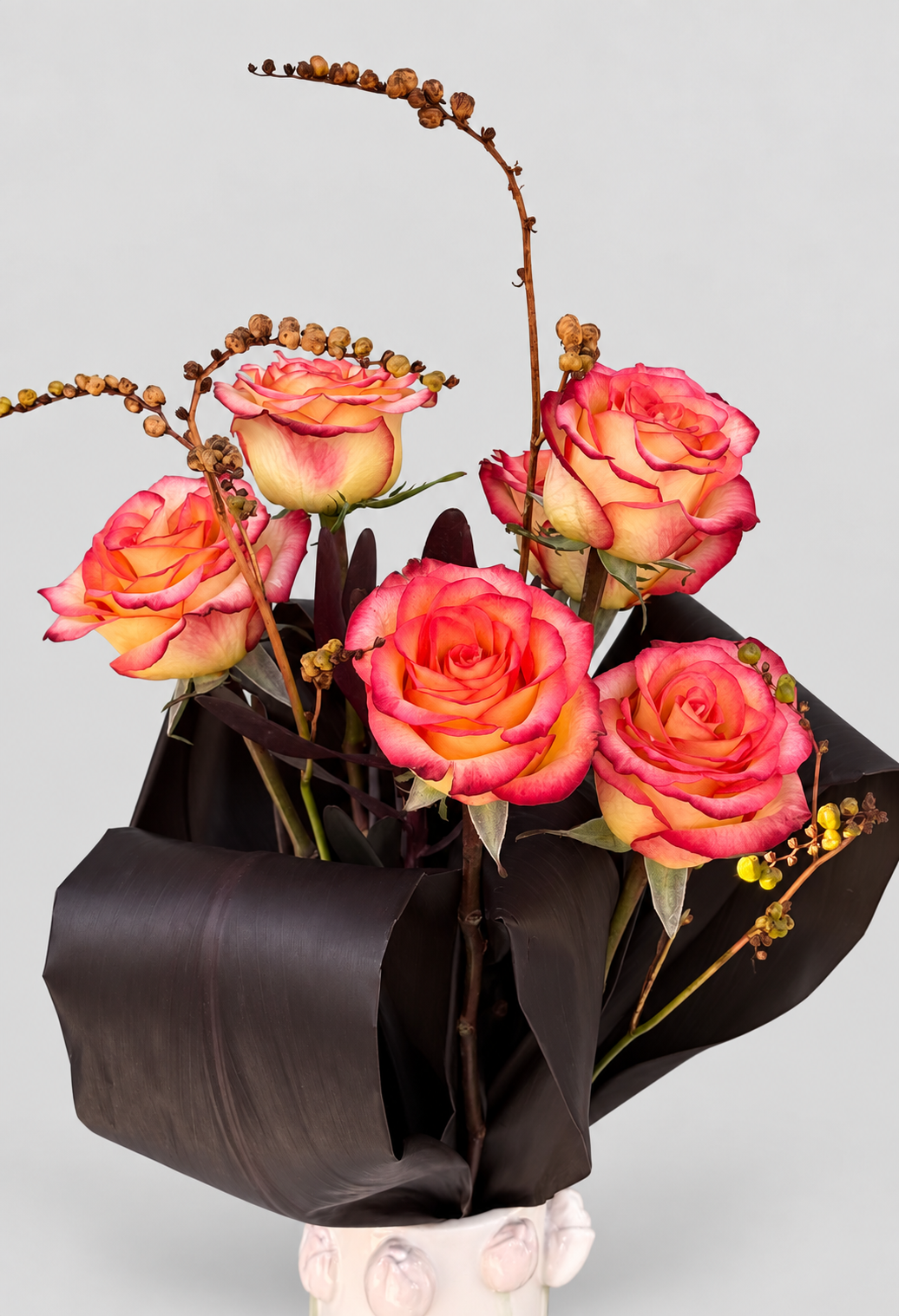 Ramo de rosas bicolor en tonos coral y amarillo con ramas de crocosmia y hojas oscuras en jarrón de cerámica blanca