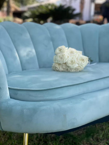 Sofá de terciopelo azul claro con respaldo curvo y ramo de rosas blancas sobre el asiento
